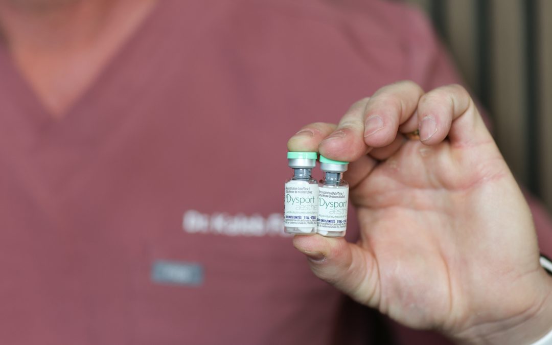 Close-up of a Kamloops cosmetic injector holding two Dysport (botulinum toxin) vials, with a blurred clinical background.
