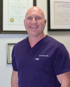 Smiling bald doctor in purple scrubs with name Dr. Kaleb Falk, standing in a clinic with framed certificates in the background
