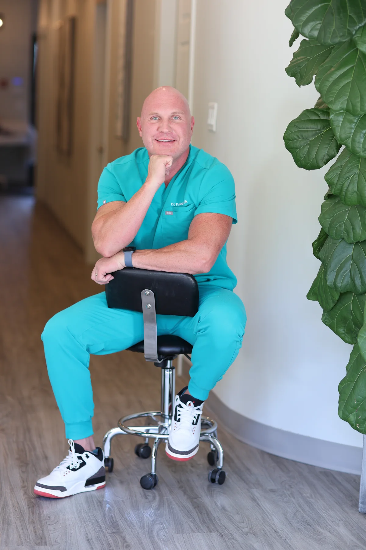 Dr. Kaleb Falk, ND, seated in turquoise scrubs beside a leafy plant.