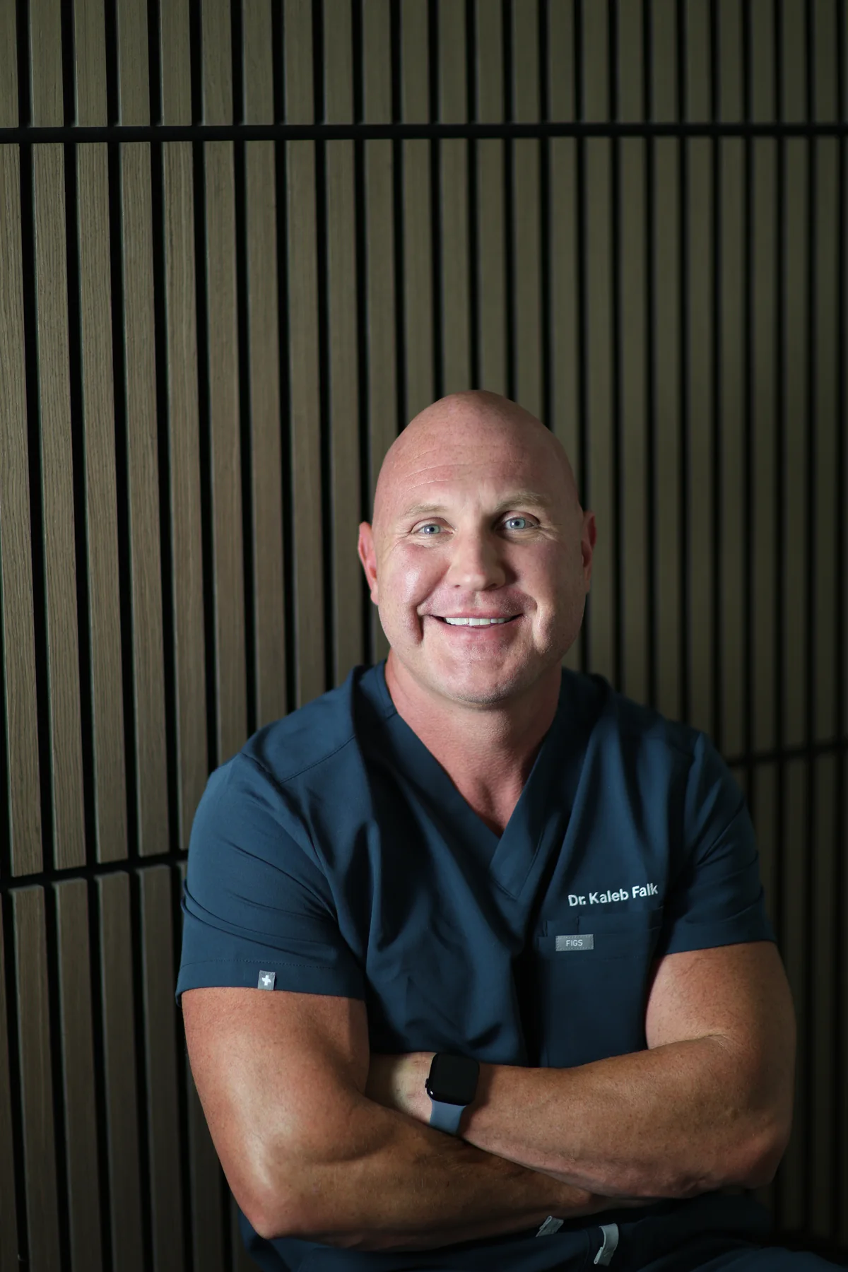 Dr. Kaleb Falk, ND, smiling in navy scrubs with his arms crossed.